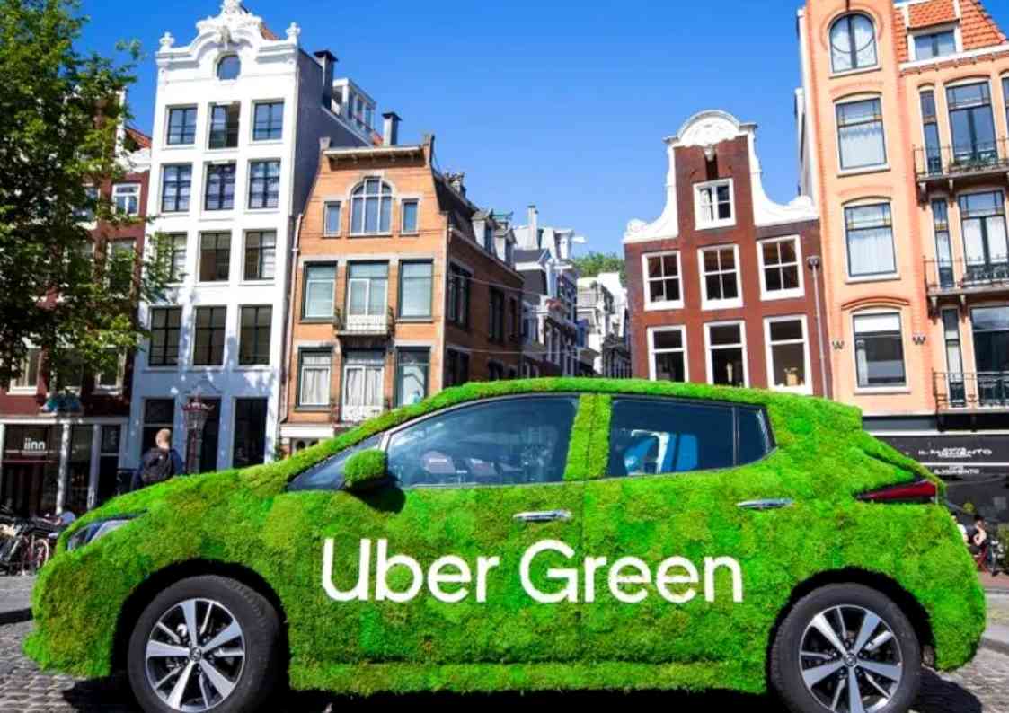Carro da Uber Green coberto com grama artificial estacionado em uma rua de uma cidade europeia, com prédios coloridos ao fundo.