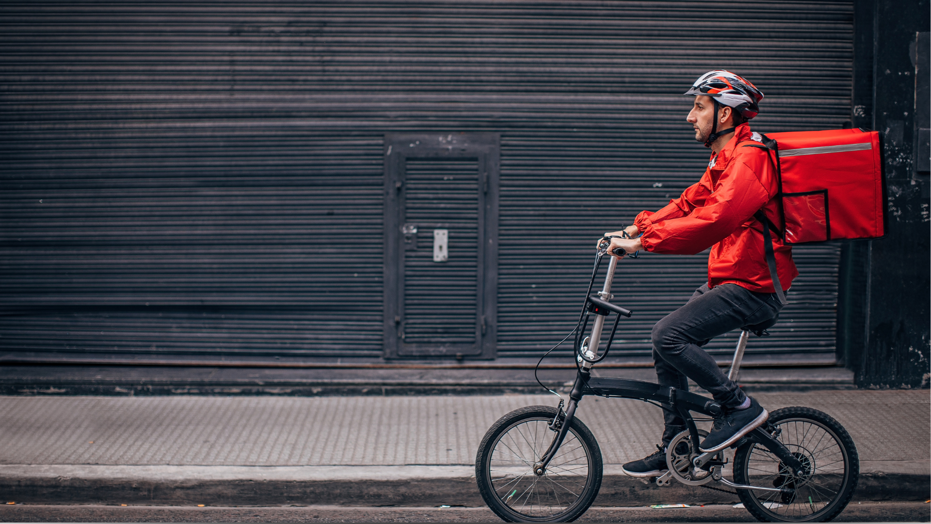 Entregador de bicicleta com mochila vermelha pedalando em uma rua urbana.