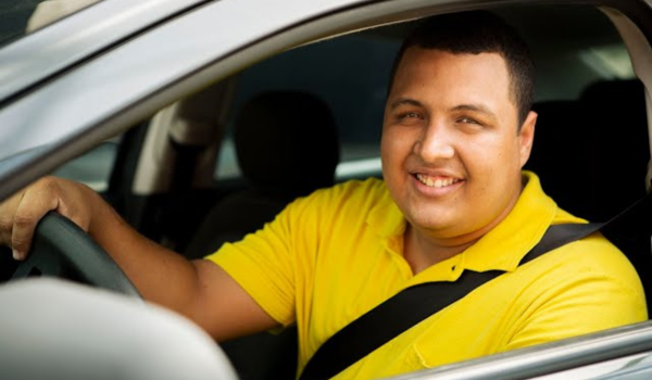 A imagem mostra um homem sorridente ao volante de um carro. Ele está usando uma camisa polo amarela e está com o cinto de segurança afivelado. O homem está olhando para a câmera, parecendo feliz ou satisfeito