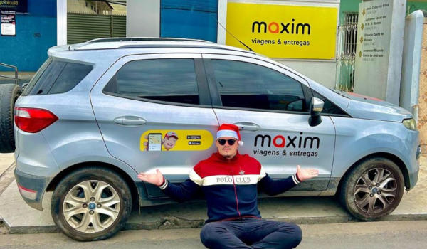 Um homem está sentado na calçada com as pernas cruzadas, à frente de um carro hatchback prateado. Ele usa um gorro de Papai Noel vermelho, óculos escuros pretos, uma camiseta branca com um logotipo no peito e um agasalho preto com listras vermelhas e brancas nas mangas. O carro tem a inscrição "maxim viagens e entregas" ao lado, junto com logotipos da marca e a ilustração de um rosto masculino. Há um edifício amarelo ao fundo com uma placa que também diz "maxim viagens e entregas".