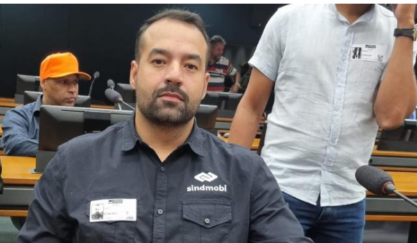 Um homem com barba leve e camisa escura com o logotipo "SINDMOBI" está sentado em uma sala de conferências, atento e olhando para a câmera, com outras pessoas ao fundo.