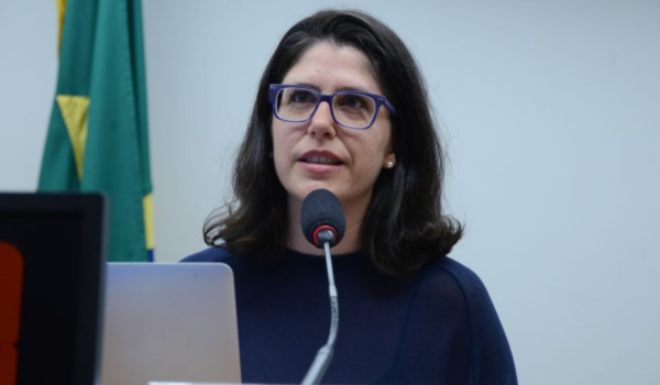 Mulher em discurso público, usando óculos de armação azul, falando ao microfone diante de um laptop. Ela tem cabelos castanhos médios, usa blusa azul escura e está em um ambiente interno com a bandeira do Brasil ao fundo.
