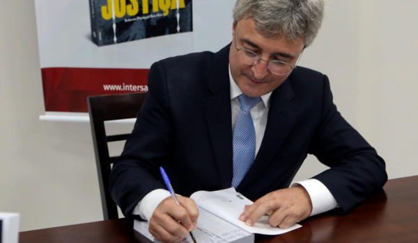 A imagem mostra um homem usando um terno e gravata, com cabelos grisalhos e óculos, sentado à mesa enquanto assina um documento. Ele parece estar concentrado na sua escrita e aponta para o local no papel onde está escrevendo.