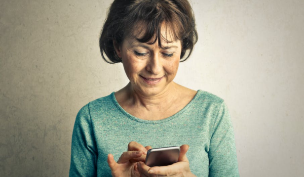 Uma mulher de meia-idade com cabelos castanhos curtos, sorrindo suavemente enquanto olha para baixo em direção a seu smartphone, que ela está usando com ambas as mãos. Ela está vestindo uma blusa azul-petróleo e está em pé contra um fundo neutro texturizado.