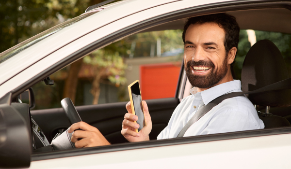 Um homem com barba e cabelos escuros, vestindo uma camisa branca e suspensórios pretos, sorri enquanto segura um smartphone com a mão direita e segura o volante de um carro com a esquerda. Ele parece estar estacionado e feliz, possivelmente usando o aplicativo no telefone. O ambiente é diurno e parece ser uma área residencial.