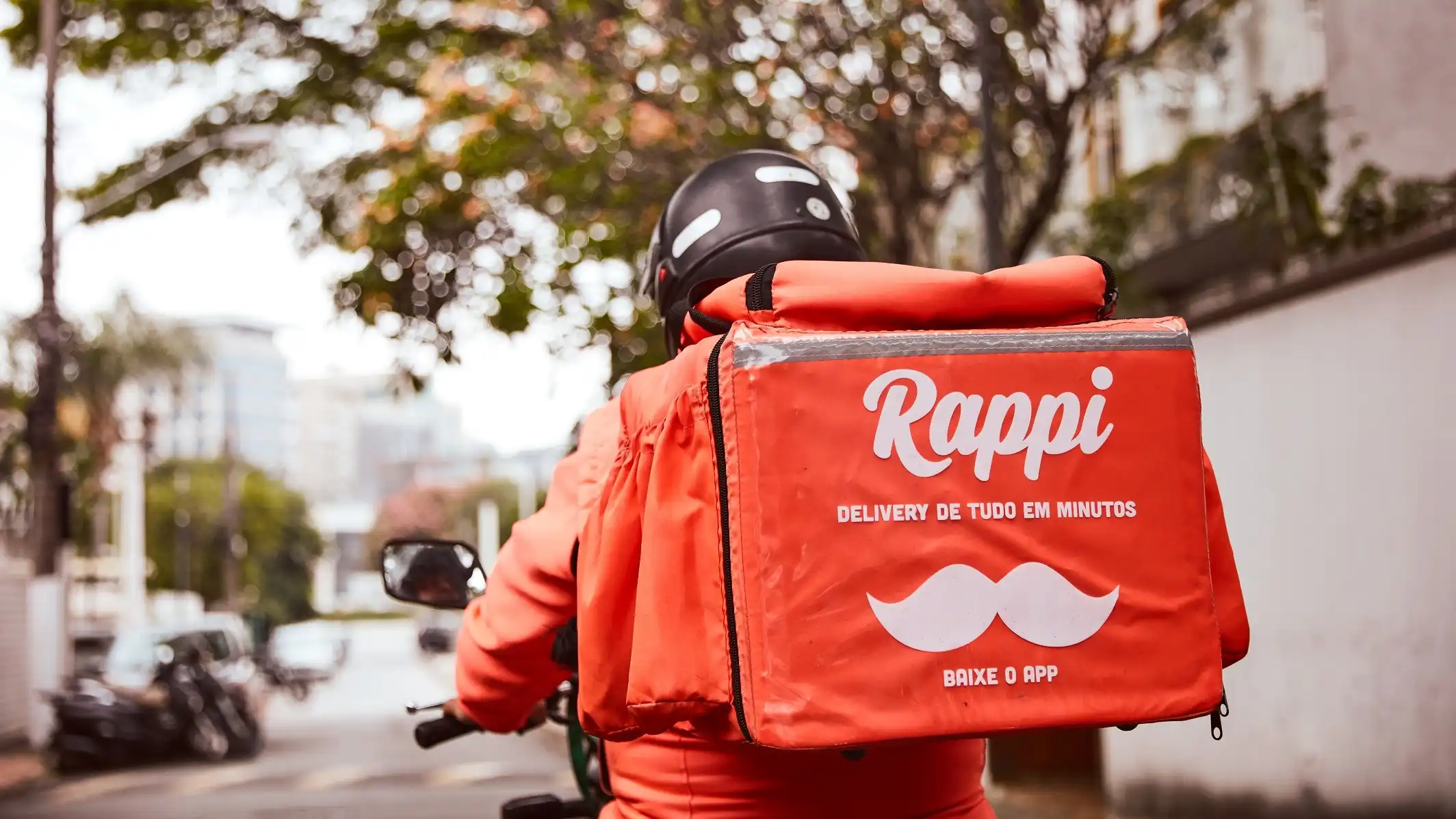 Entregador com uma mochila da Rappi laranja em uma moto.