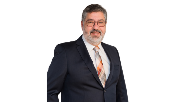 Retrato profissional de um homem sorridente com cabelo grisalho, barba e bigode. Ele está usando óculos, um terno escuro, camisa branca e uma gravata estampada em tons de laranja e azul.