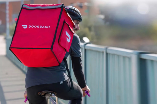 Entregador da DoorDash pedalando uma bicicleta em uma ponte, vestindo uma mochila térmica vermelha com o logo da DoorDash nas costas. O entregador usa um capacete preto e está de costas para a câmera, enquanto entrega um pedido.