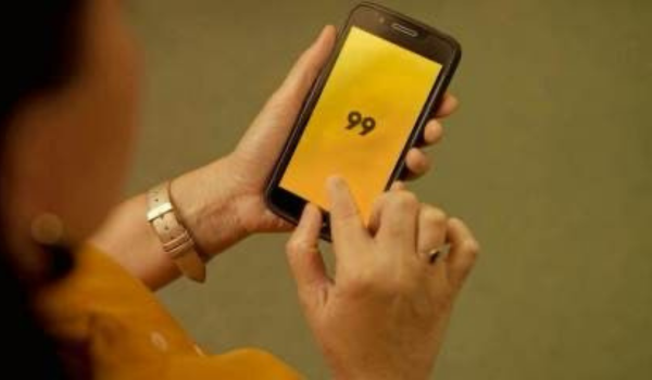 Pessoa segurando um smartphone com a tela voltada para cima, exibindo um fundo amarelo brilhante com o número '99' em grande destaque no centro. A pessoa está usando uma blusa laranja e tem um relógio de prata no pulso esquerdo. Somente a mão e o antebraço da pessoa estão visíveis na imagem.