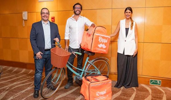 Três pessoas sorridentes em pé ao lado de uma bicicleta laranja com uma cesta e uma caixa térmica também laranja. Um homem à esquerda veste óculos, camisa social e calças, segurando um capacete de bicicleta. O homem ao centro, de camiseta e calça jeans, apoia-se na bicicleta, que tem o logo de uma empresa de entrega. Uma mulher à direita, de blazer branco e calça preta, com cabelo longo, completa o grupo. Estão em uma sala com carpete e uma saída de emergência ao fundo.