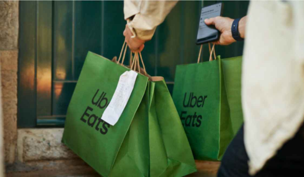 A imagem apresenta uma pessoa deixando sacolas verdes em uma porta. Nas sacolas está escrito em preto "Uber Eats", indicando ser uma possível entrega da empresa.