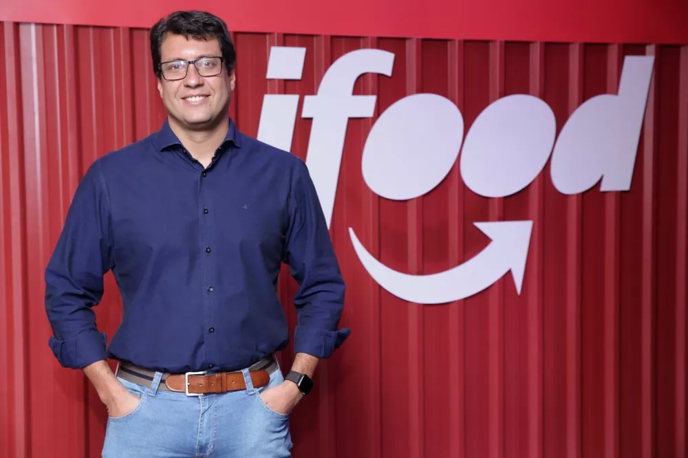 "Um homem sorridente em uma camisa azul escura e calça jeans está em pé diante de um fundo com o logotipo do iFood, que é predominantemente vermelho com o nome da marca em branco."