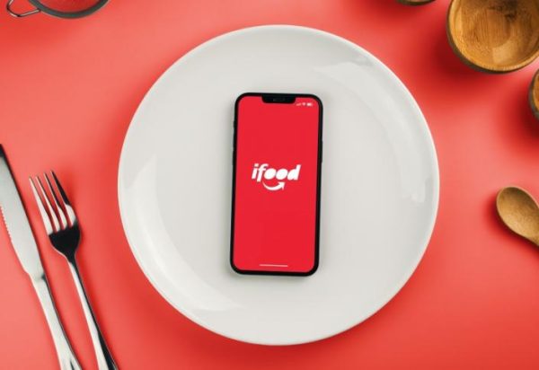 A imagem mostra um smartphone com a tela exibindo o logo do iFood, posicionado no centro de um prato branco. O prato está sobre uma superfície vermelha, e ao redor dele há talheres e uma xícara, sugerindo um cenário de refeição.