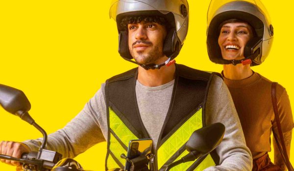 A imagem mostra duas pessoas sorridentes em um fundo amarelo vibrante. Elas estão vestindo capacetes de segurança para andar de moto, com o homem à frente segurando o guidão de uma motocicleta e a mulher atrás dele, sugerindo que são passageiros. O homem usa uma camiseta de manga comprida cinza e um colete refletor verde fluorescente. Ele está olhando para a frente, com uma expressão confiante. A mulher atrás dele veste uma blusa de manga comprida marrom e também um capacete, sorrindo levemente e parecendo estar aproveitando o passeio. Ambos têm um estilo casual e urbano, e parecem estar preparados para uma viagem de moto segura.