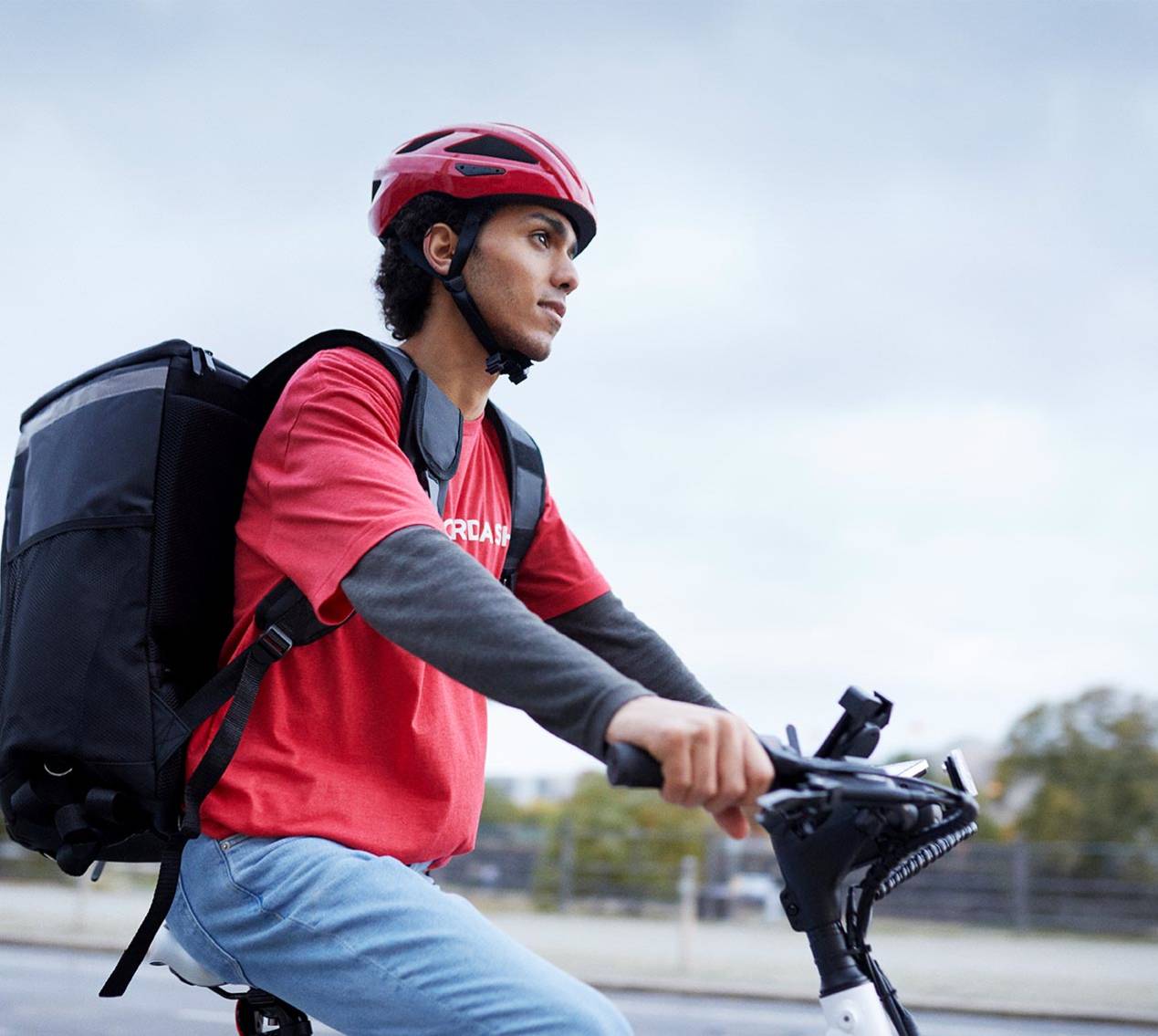 A imagem mostra um entregador usando um capacete vermelho, uma camiseta vermelha e uma mochila de entrega preta nas costas enquanto anda de bicicleta. Ele parece focado e está pedalando em uma área urbana, provavelmente realizando uma entrega. A cena sugere um dia de trabalho típico para entregadores de serviços de entrega rápida.