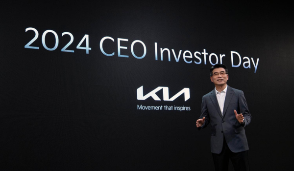Homem de negócios asiático de meia-idade apresentando-se em um evento, diante de um fundo preto com texto branco que diz '2024 CEO Investor Day'. Ao lado do texto está o logotipo da Kia Motors, uma marca estilizada em branco com o slogan 'Movement that inspires' abaixo.