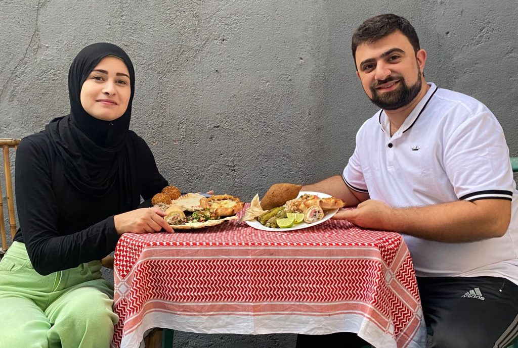 A imagem mostra um homem e uma mulher sentados à mesa, ambos sorrindo para a câmera enquanto se preparam para comer. A mulher, vestindo hijab preto e roupas casuais verdes, está à esquerda. O homem, à direita, veste uma camisa polo branca e calças esportivas. Eles estão compartilhando uma refeição de pratos do Oriente Médio, como falafel, kebab e saladas, servidos em uma mesa coberta com uma toalha xadrez vermelha e branca. O fundo é uma parede de concreto cinza.