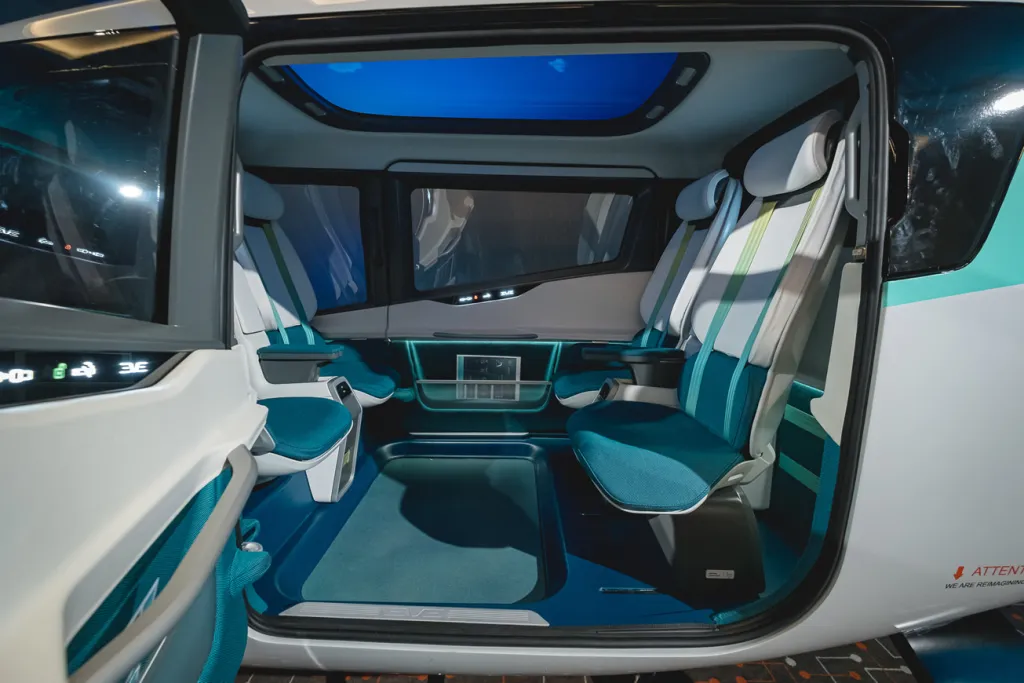 "Interior de uma aeronave eVTOL da Eve Air Mobility, mostrando quatro assentos dispostos em configuração de face a face, com estofamento em tecido azul-turquesa e detalhes em branco e verde. O espaço é moderno e minimalista, com janelas amplas para vistas externas, painéis de controle integrados nas laterais da cabine e iluminação ambiente em tom azul suave. O logotipo da Embraer e a frase 'We are reimagining flight' são visíveis no limiar da porta aberta."