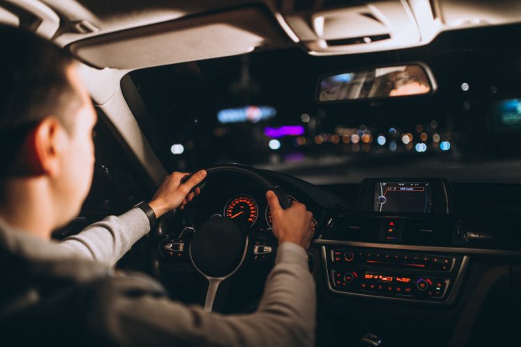 Homem dirigindo seu carro à noite, com o painel iluminado e a estrada à frente visível através do para-brisa.