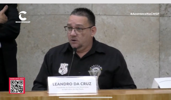 Um homem de meia-idade, usando óculos e camisa preta com um emblema no peito esquerdo, está falando ao microfone durante um evento público. Ele está identificado na placa em frente a ele como "LEANDRO DA CRUZ", Presidente do Sindicato dos Motoristas de Aplicativo de São Paulo. O cenário inclui um fundo cinza claro e uma mesa de madeira. No canto inferior esquerdo, há um QR code com o texto "SAIBA MAIS AQUI", indicando informações adicionais disponíveis para o espectador.