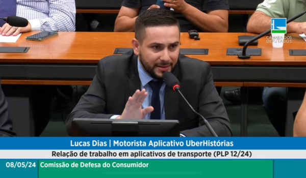 Lucas Dias, um motorista de aplicativo, participa de uma comissão na Câmara dos Deputados. Ele está falando ao microfone, vestido com um terno azul e gravata, enquanto gesticula com a mão direita. No fundo, vê-se outras pessoas sentadas ouvindo. Informações na tela incluem texto sobre a discussão de legislação relacionada a trabalhos em aplicativos de transporte.