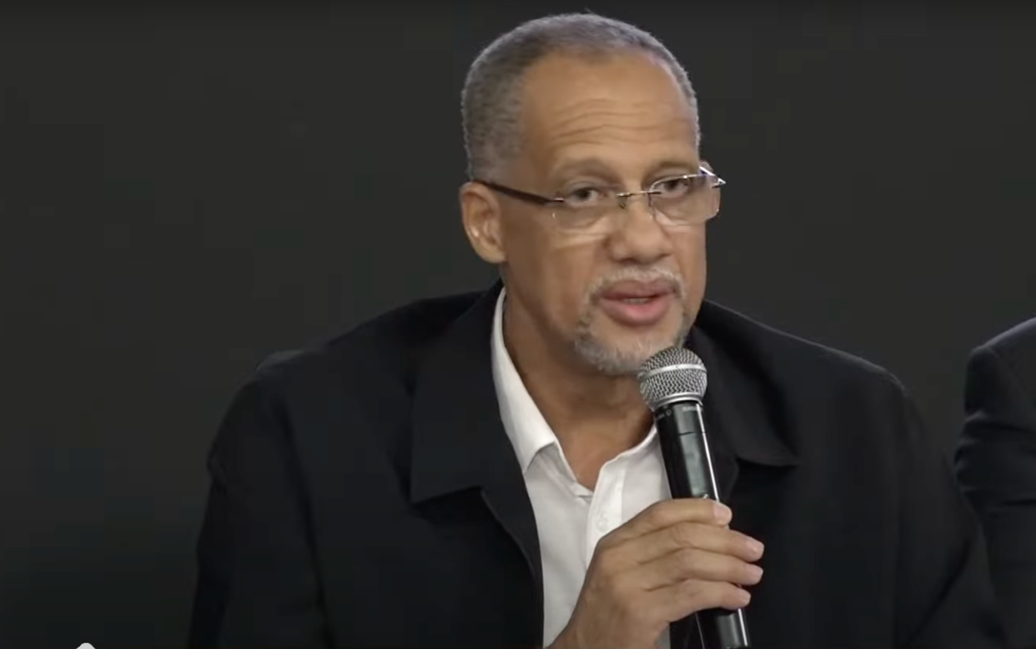 Cláudio Sena, homem de óculos, usando terno preto e camisa branca, segurando um microfone e falando em um evento.