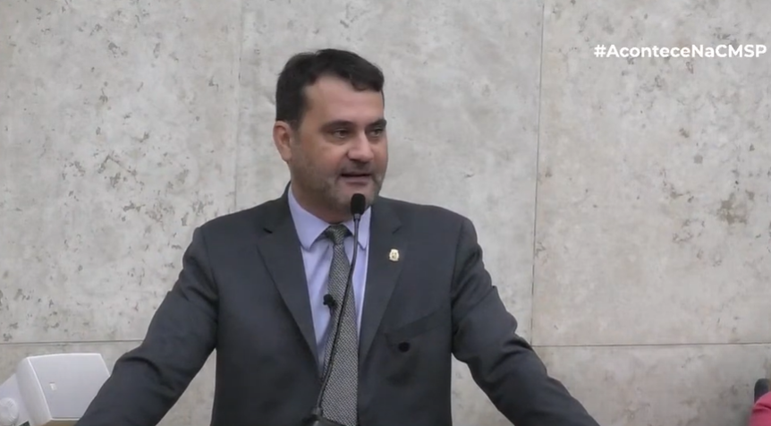 Homem de terno discursando em púlpito com microfone durante sessão da Câmara Municipal de São Paulo.