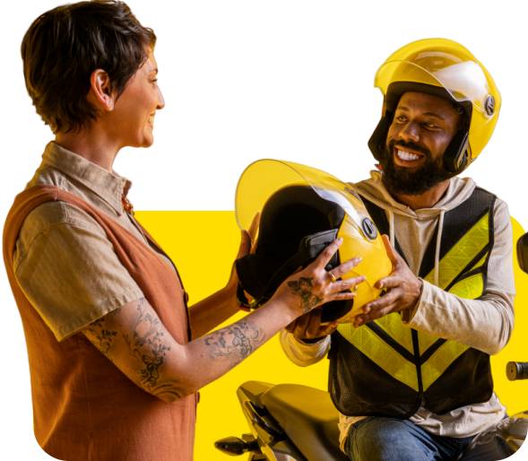 Uma mulher e um homem estão parados ao lado de uma motocicleta amarela. A mulher está entregando um capacete amarelo para o homem, que está sorrindo. Ambos estão usando coletes de segurança, e há um fundo amarelo atrás deles.