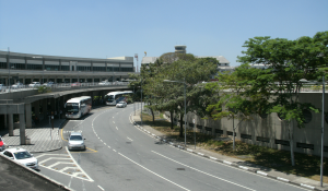 Vista externa de um terminal de aeroporto com uma rua curva em frente. Há alguns carros e ônibus estacionados ao lado do terminal, árvores ao longo da via, e um céu claro sem nuvens. A estrutura do aeroporto é extensa, com um prédio circular ao fundo.