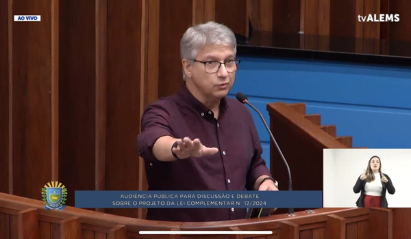 Um homem de cabelos grisalhos e óculos está falando em um púlpito durante uma audiência pública. Ele veste uma camisa de manga longa marrom e está com uma das mãos levantada em um gesto de ênfase. No canto inferior direito, há uma intérprete de Libras (Língua Brasileira de Sinais) gesticulando. No rodapé da imagem, há uma faixa azul com o texto: "Audiência Pública para Discussão e Debate sobre o Projeto da Lei Complementar nº 12/2024". O logotipo da tvALEMS está no canto superior direito.