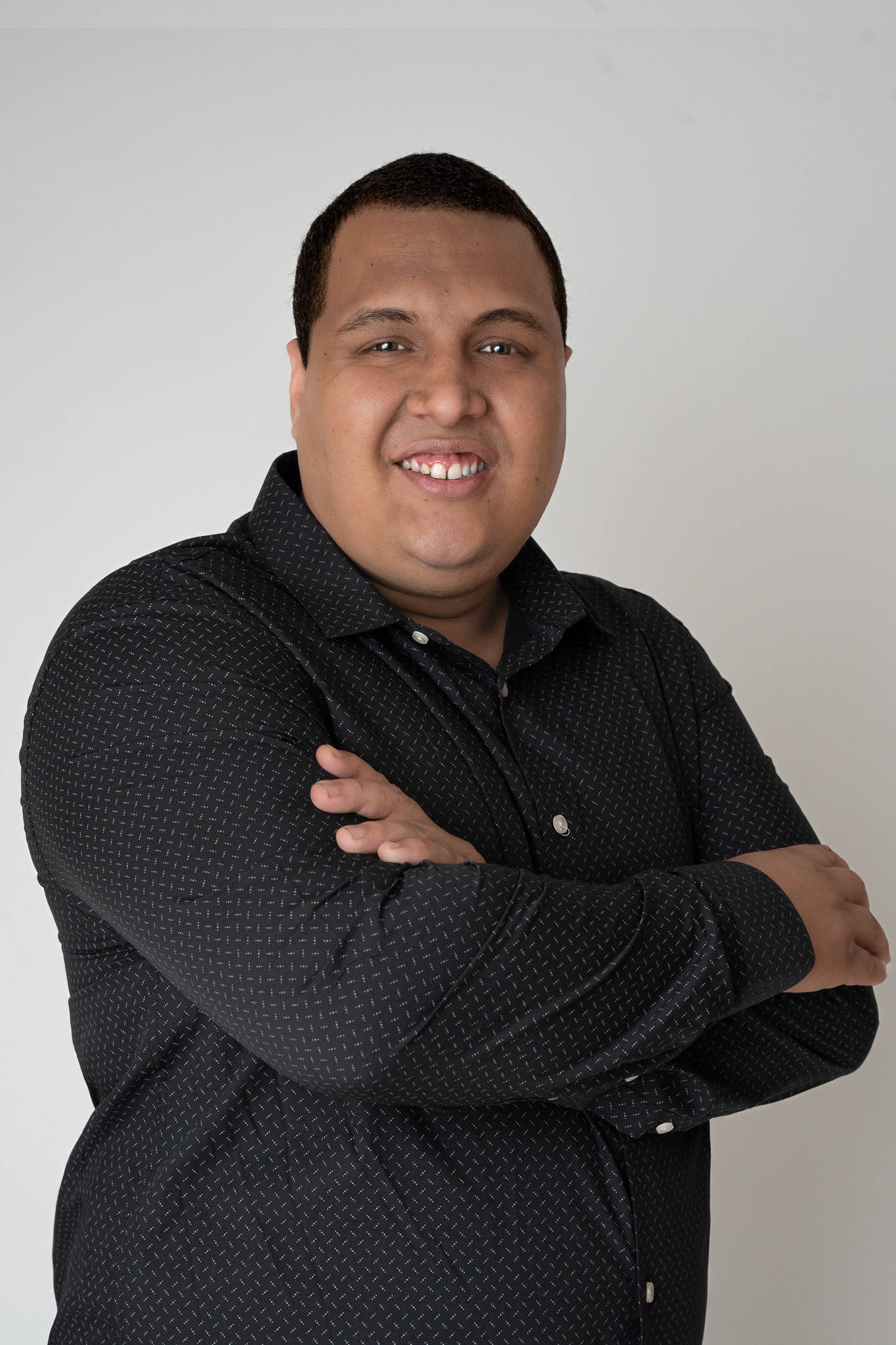 Um homem sorridente de braços cruzados, vestindo uma camisa preta de mangas compridas com um padrão discreto. Ele tem cabelo curto e está posicionado contra um fundo branco simples.