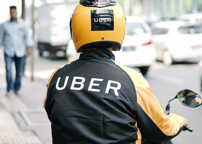 Entregador do Uber Flash Moto em motocicleta, usando jaqueta e capacete da Uber em uma rua movimentada.