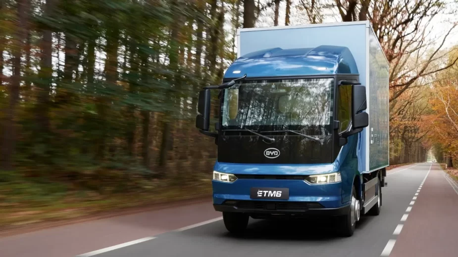 Um caminhão elétrico azul, modelo ETM6 da marca BYD, está em movimento em uma estrada cercada por árvores em um cenário de outono. O veículo ocupa a maior parte da imagem, com detalhes claros da frente e da lateral do caminhão, enquanto a estrada e a vegetação ao redor passam rapidamente, sugerindo que o caminhão está em alta velocidade.
