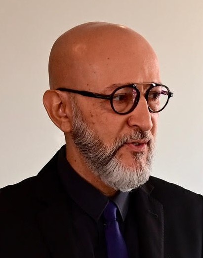 A imagem mostra um homem careca, com barba e óculos de armação redonda.