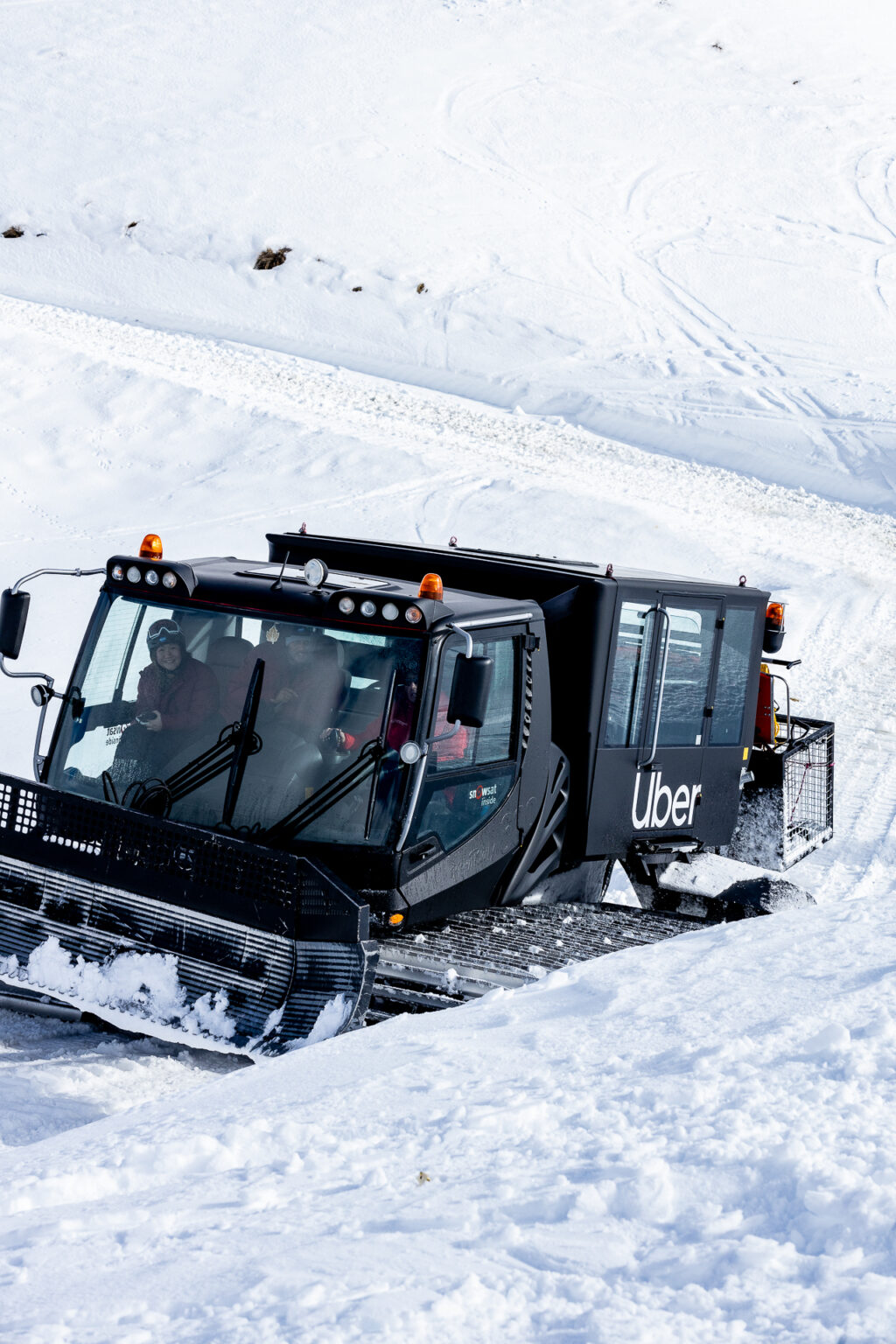 A imagem mostra um veículo tipo snowcat, adaptado para operar em áreas com neve, com a marca "Uber" visível em sua lateral. O snowcat está em movimento sobre uma superfície coberta de neve, com duas pessoas visíveis dentro da cabine do veículo. O cenário ao redor é completamente nevado, sugerindo um ambiente de montanha ou estação de esqui. O veículo parece ser utilizado para transporte em terrenos nevados, como parte de uma iniciativa ou serviço oferecido pela Uber em locais com neve abundante.