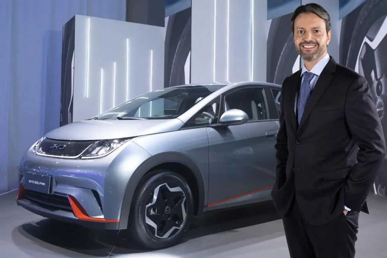A imagem mostra um homem de terno e gravata, sorrindo, em pé ao lado de um carro elétrico prateado da marca BYD, modelo Dolphin. Ao fundo, há um cenário moderno com luzes verticais brilhantes, destacando o ambiente tecnológico e inovador da apresentação.