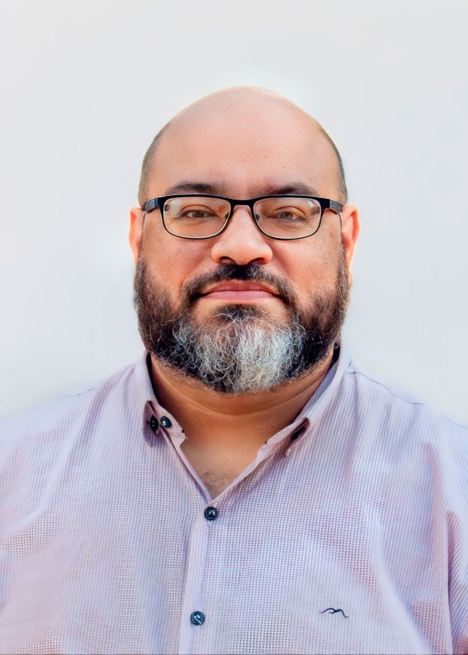 Uma foto de André Gomide, um homem barbudo com óculos e cabeça calva, vestindo uma camisa clara com botões. Ele parece estar em pé diante de um fundo claro e neutro, olhando diretamente para a câmera com uma expressão neutra.