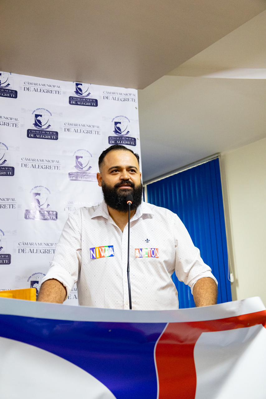 Marcelo Alves, pré-candidato a vereador em Alegrete, fala ao público em um evento. Ele está de pé atrás de um pódio, vestindo uma camisa branca com os nomes "Nival" e "Antion" colados no peito. No fundo, há um banner da Câmara Municipal de Alegrete. Marcelo tem barba e cabelo curto, e parece estar discursando com um microfone à sua frente.