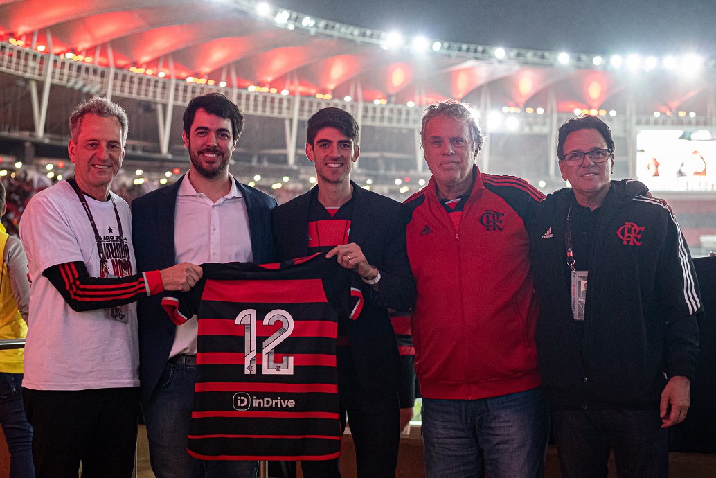 A imagem mostra cinco homens posando juntos no estádio do Maracanã. Eles estão segurando uma camisa do Flamengo com o número 12 e o logo da inDrive, indicando uma parceria entre o clube e o aplicativo de transporte. A iluminação vermelha do estádio ao fundo e o uniforme do Flamengo que alguns deles estão vestindo ressaltam a conexão com o time de futebol.
