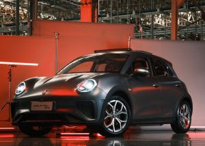 A imagem mostra um carro hatchback compacto e moderno, com carroceria cinza fosca e teto preto, em um ambiente iluminado com luzes vermelhas e brancas. O design elegante e futurista do veículo é destacado por faróis redondos com luzes diurnas LED e rodas de liga leve esportivas. O cenário parece ser um showroom ou uma área de exposição com um fundo industrial, realçando ainda mais as linhas suaves e curvilíneas do carro.
