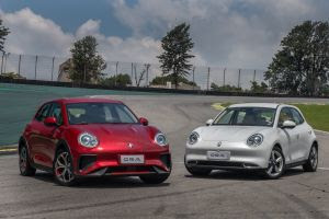 Na imagem, há dois carros elétricos da marca ORA, modelo Cat, estacionados em uma pista de corrida. O carro à esquerda é vermelho e tem um design esportivo, enquanto o carro à direita é branco e apresenta um estilo mais clássico. Ambos os carros têm faróis redondos e uma estética que mistura elementos retrô com um toque moderno. A pista ao fundo sugere um ambiente de testes ou exibição de veículos.