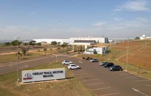 A imagem mostra a fachada da planta da Great Wall Motor Brasil, localizada em uma área ampla com bastante espaço ao redor. Em primeiro plano, há um estacionamento com alguns carros estacionados e uma placa grande indicando o nome da empresa, "Great Wall Motor Brasil". Ao fundo, é possível ver as instalações industriais da fábrica, com grandes galpões e uma estrutura moderna. O céu está claro, sugerindo um dia ensolarado.