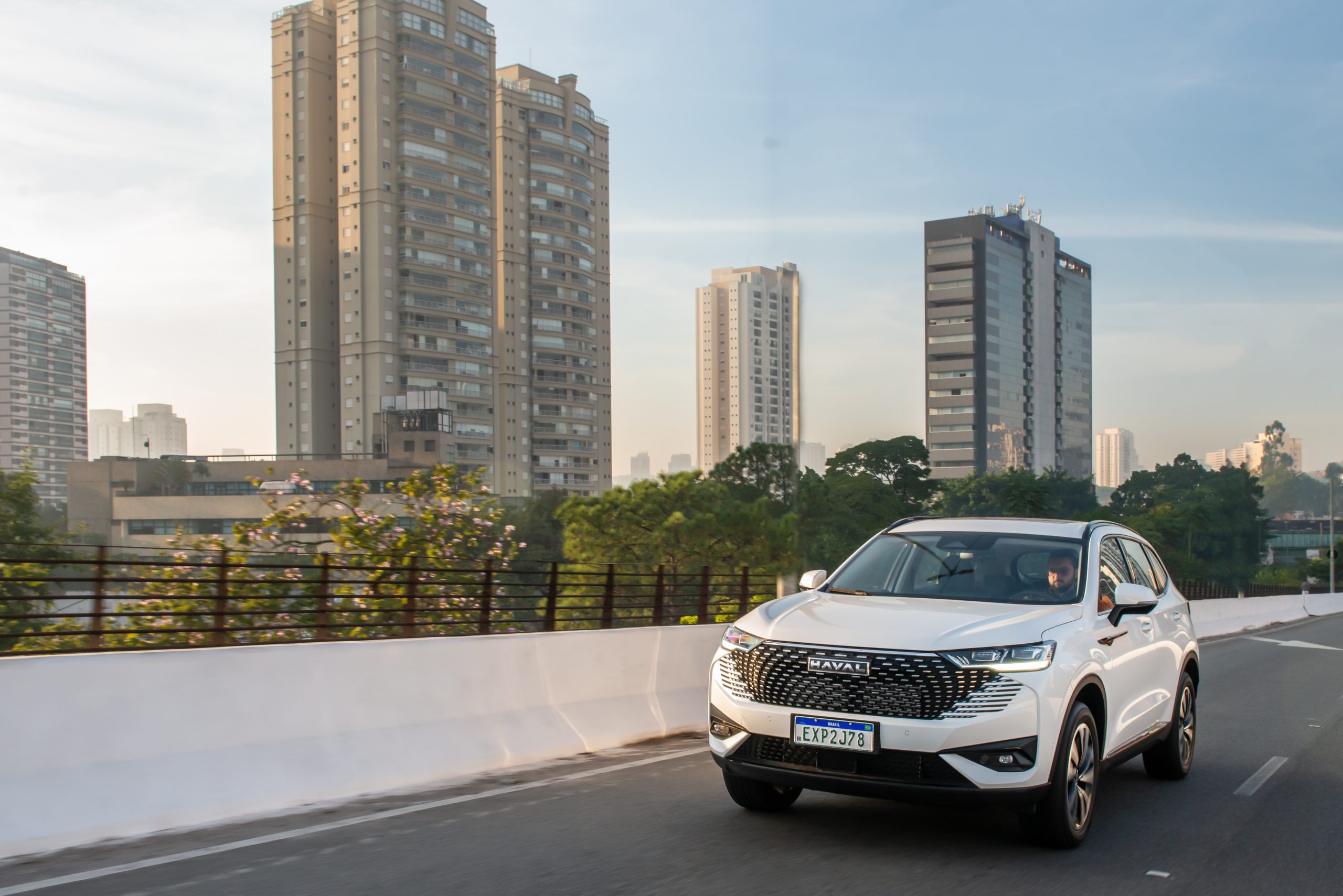 Carro SUV branco da marca Haval circulando em uma avenida de uma cidade grande, com prédios ao fundo.