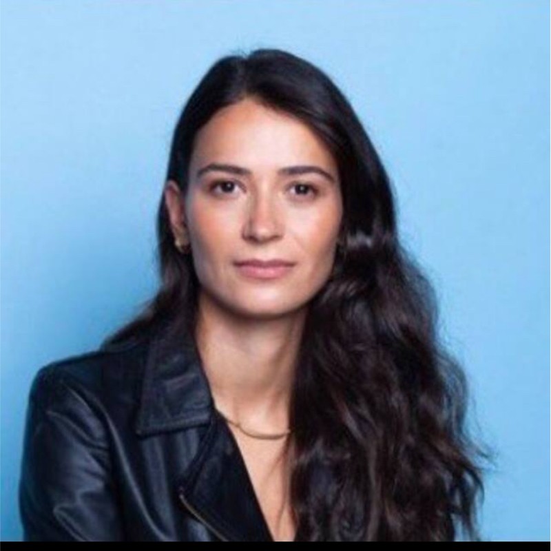 Mulher em pose formal, com cabelo solto e vestindo jaqueta preta, em fundo azul claro.