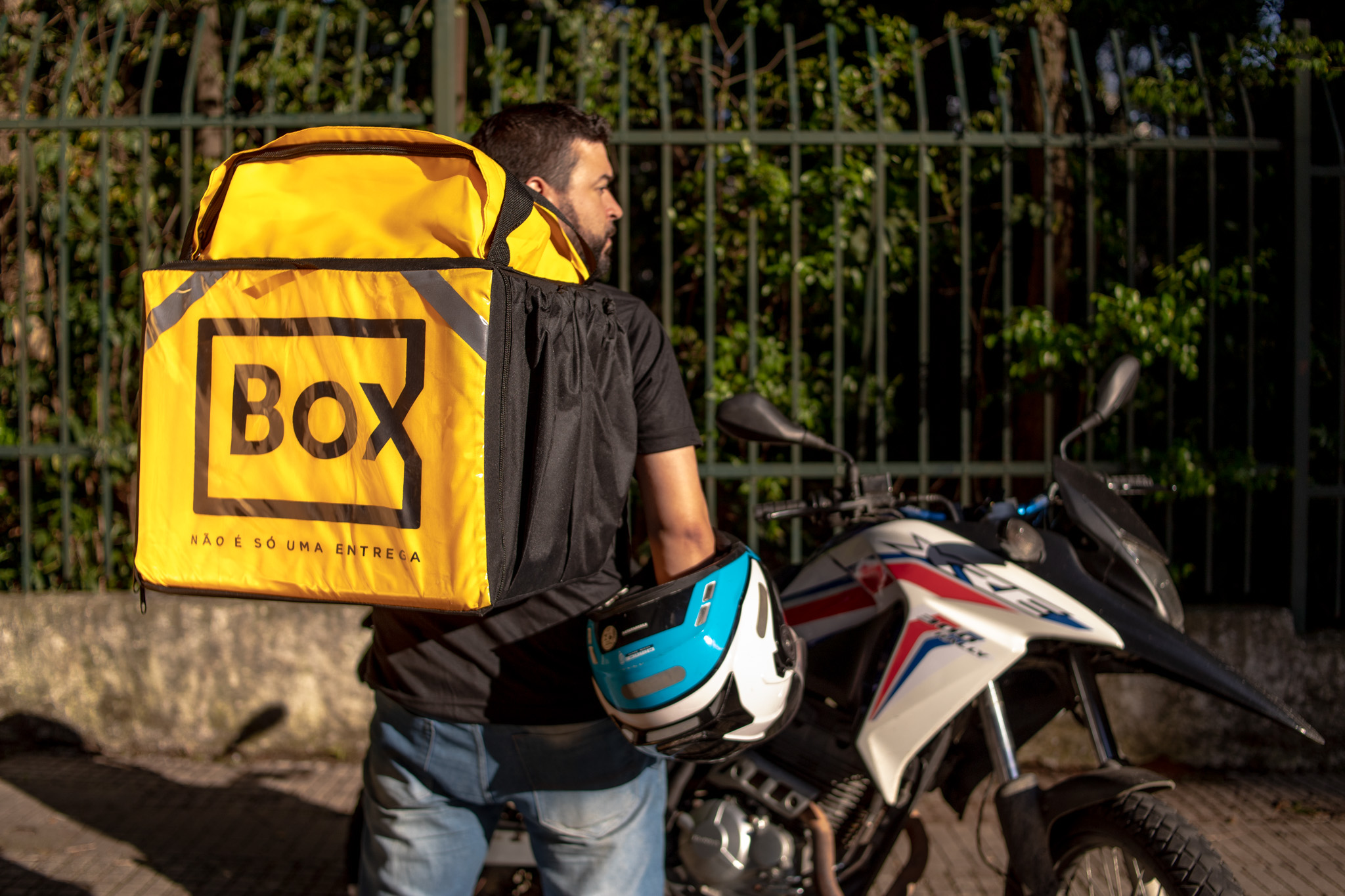 Na imagem, um entregador de moto aparece de costas, carregando uma mochila amarela Box, destacando o lema "Não é só uma entrega". Ele está segurando um capacete azul e branco ao lado de uma moto estacionada. Essa cena representa o cotidiano de entregadores de aplicativos que utilizam motos para transportar mercadorias e refeições.