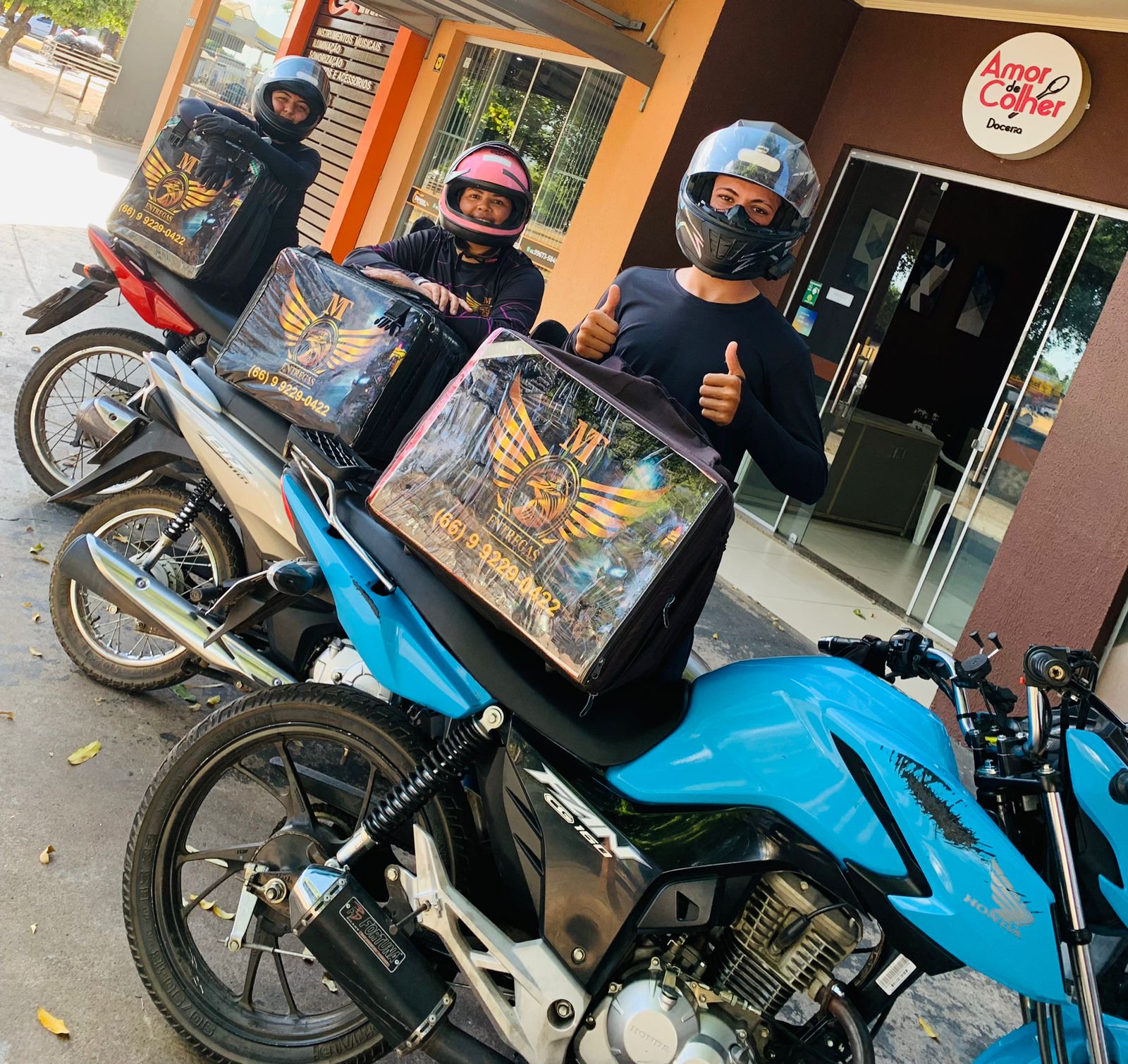 "Entregadores de moto com mochilas personalizadas de delivery posando em frente à doceria 'Amor de Colher'."