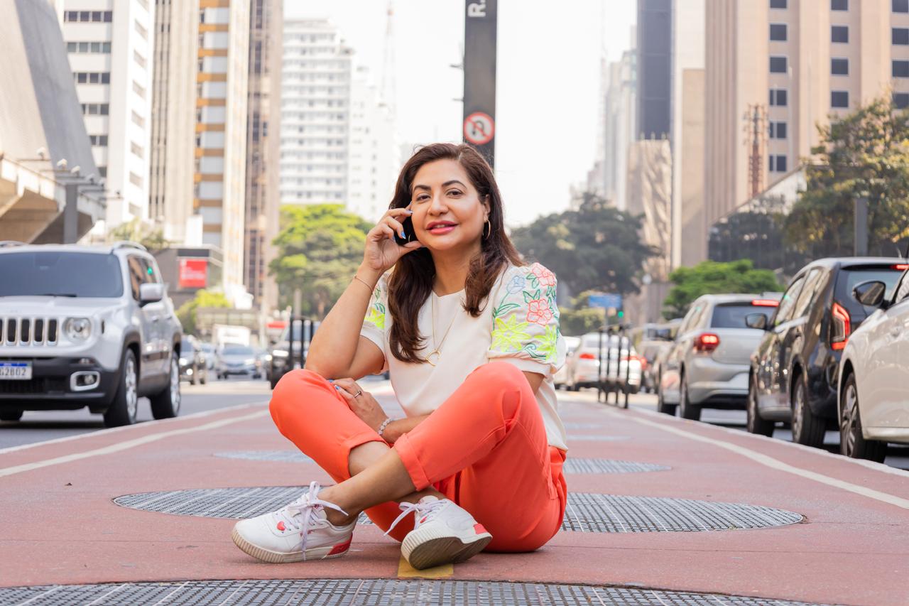 "Mulher sentada em uma avenida movimentada de São Paulo enquanto fala ao telefone, usando roupas casuais coloridas."