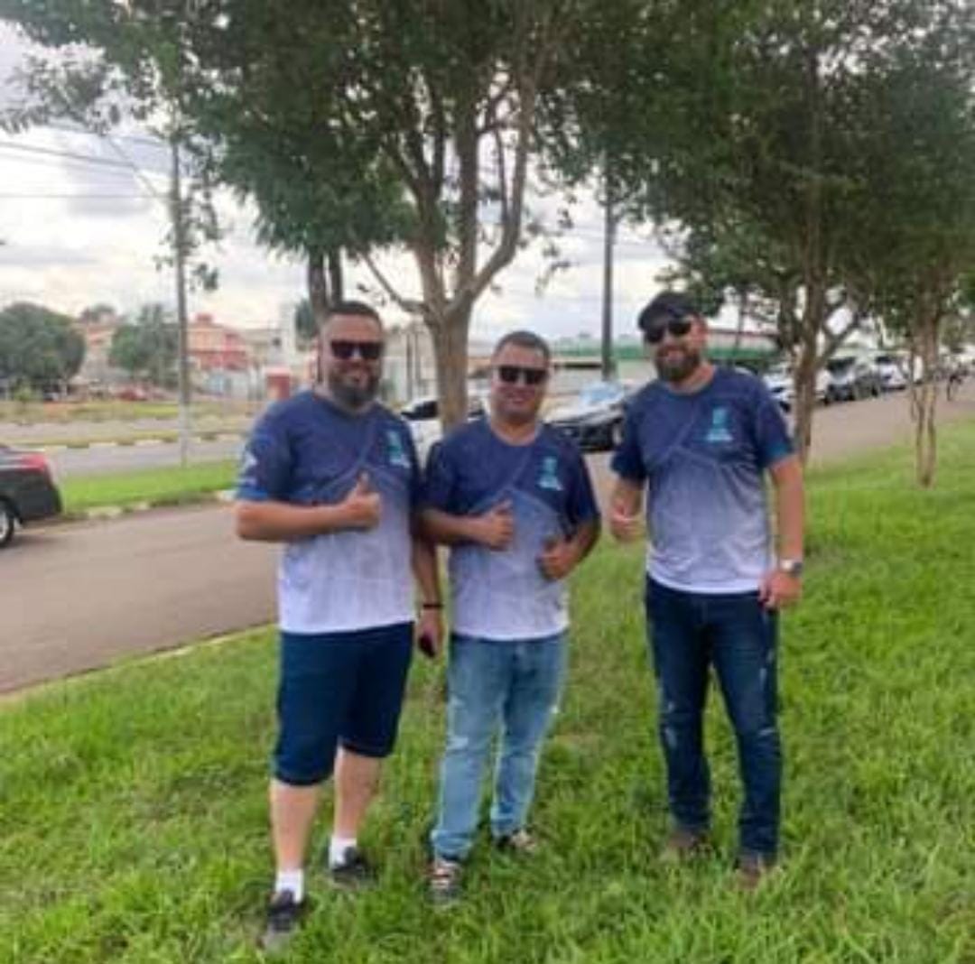 Três homens posando ao ar livre, usando camisetas azul e branca, em uma área verde.