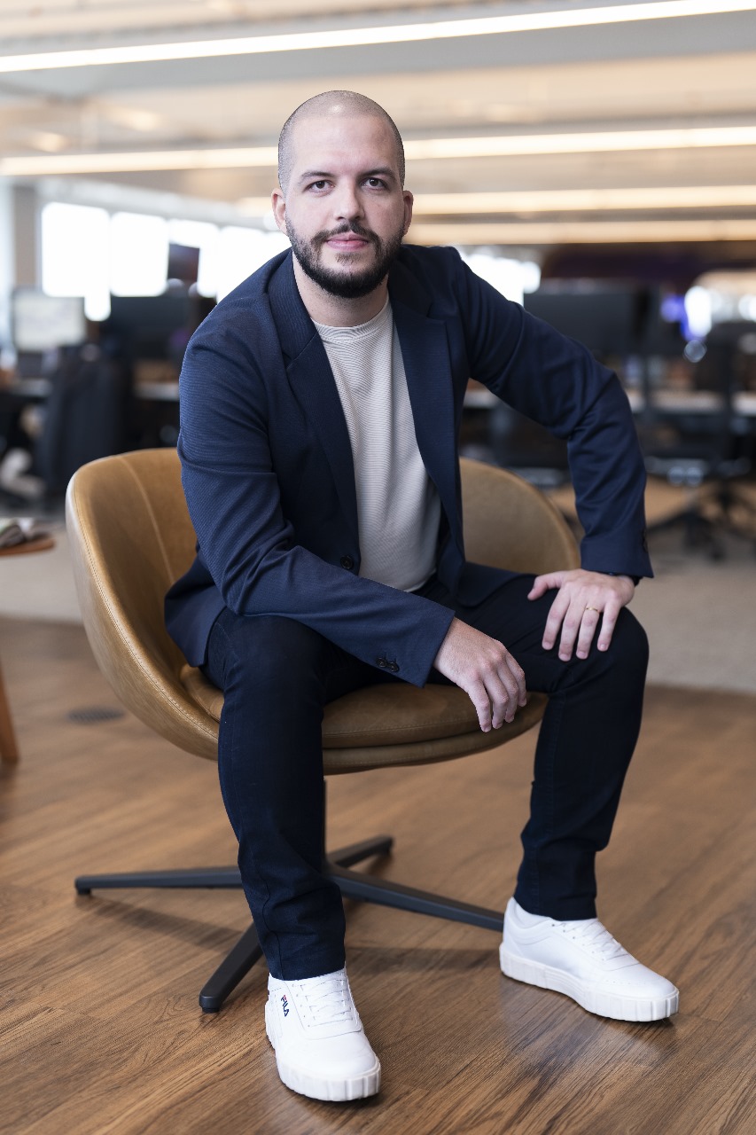 "Homem sentado em uma cadeira de escritório, usando blazer azul, calça jeans escura e tênis branco, em um ambiente corporativo moderno."