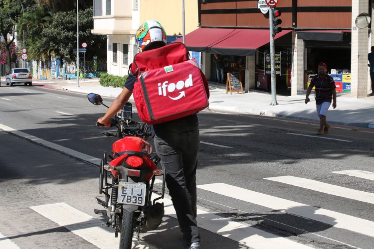 A imagem retrata um entregador do iFood com sua mochila térmica vermelha, atravessando a rua ao lado de sua motocicleta.